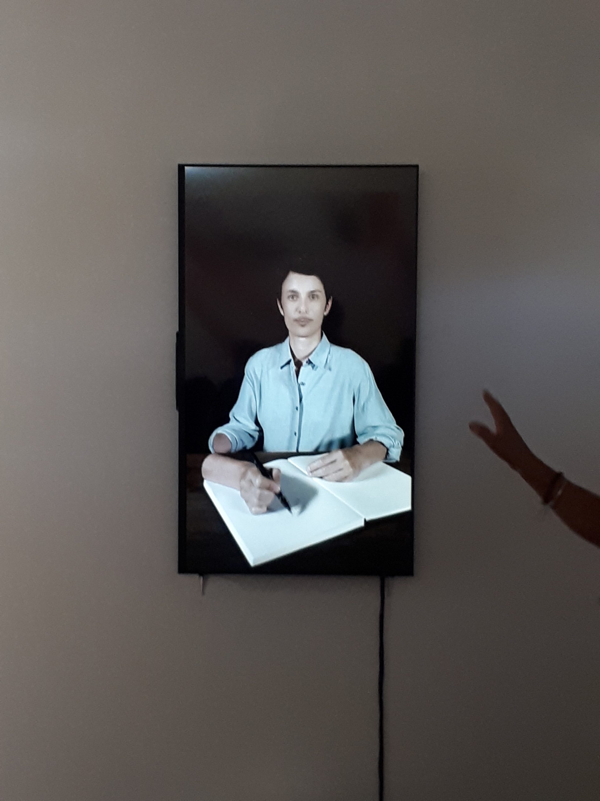 Vue d'un écan LCD grand format avec l eportrait d'un homme moustachu assis à une table. Sa main est détaché de son bras et écrit sur un papier.