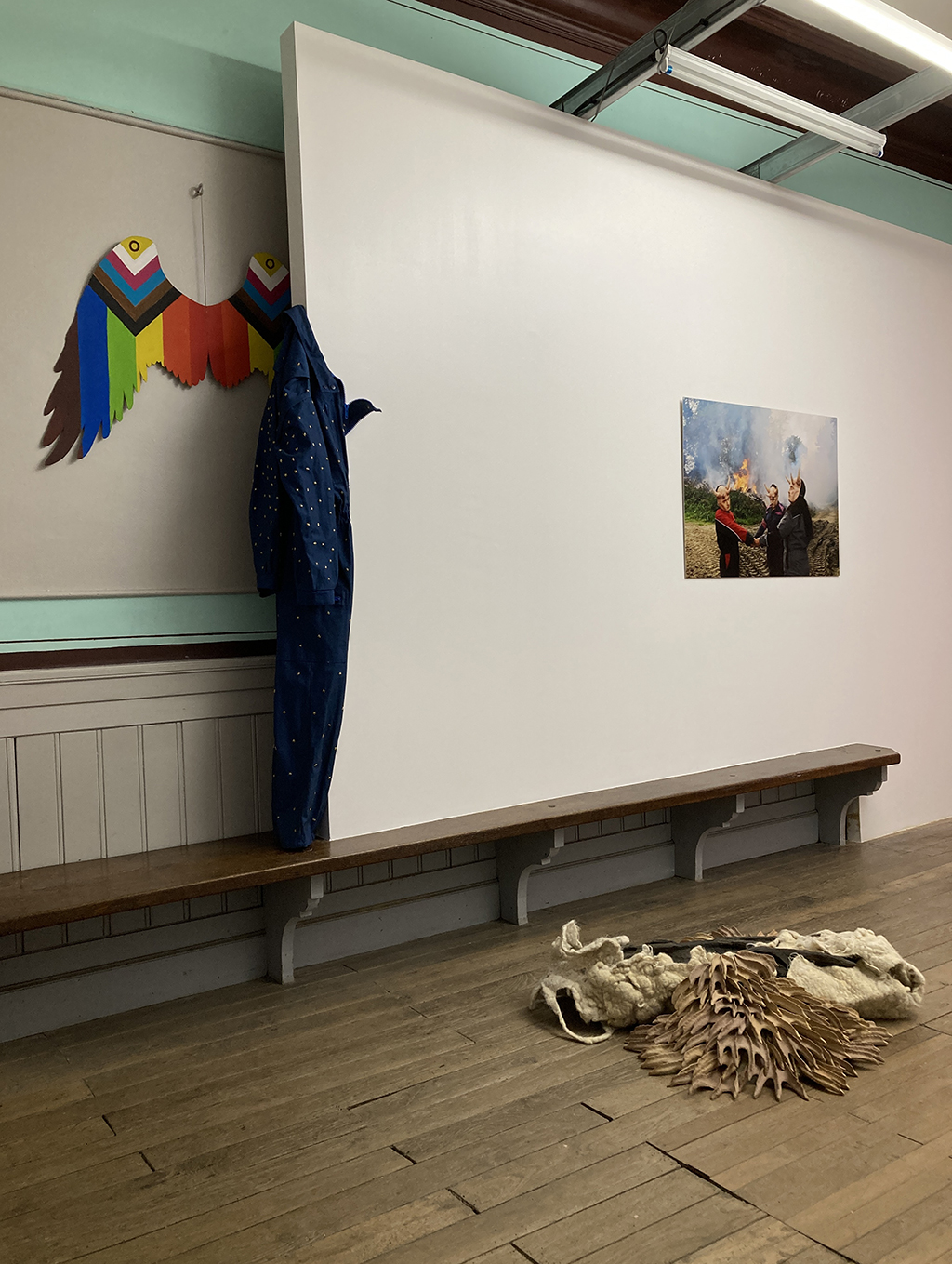 Vue de l'exposition avec une tunique d'agriculteur cloutée d'étoile, des ailes en carton multicolores, une sculpture en bois, laine et céramique et une photo au fond