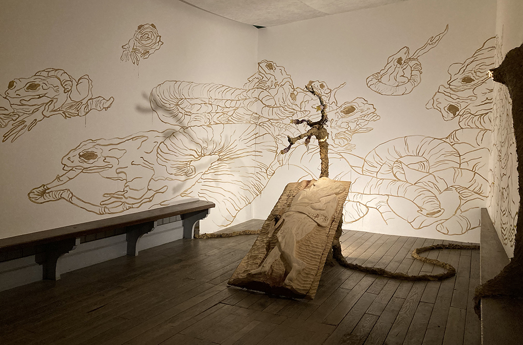 Vue de l'exposition avec une fresque réalisée avec su jus de terre, représentant des grenouilles et des sangsues, et une sculpture en bois d'un gisant tenant un bouquet de blé.